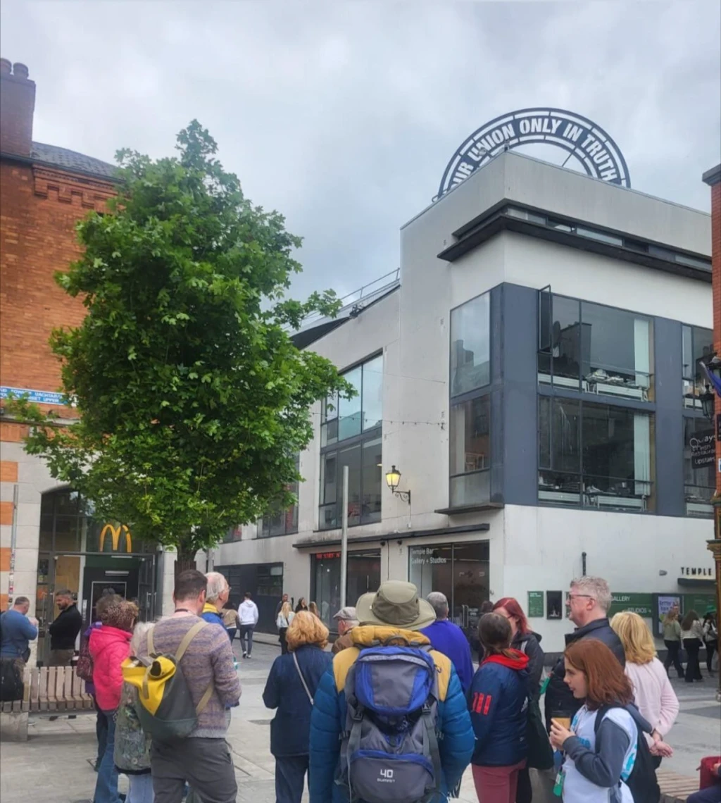 A group of people gathered in a square lined by concrete and red brick buildings. There is a bright green tree in the square, and the sky is cloudy.