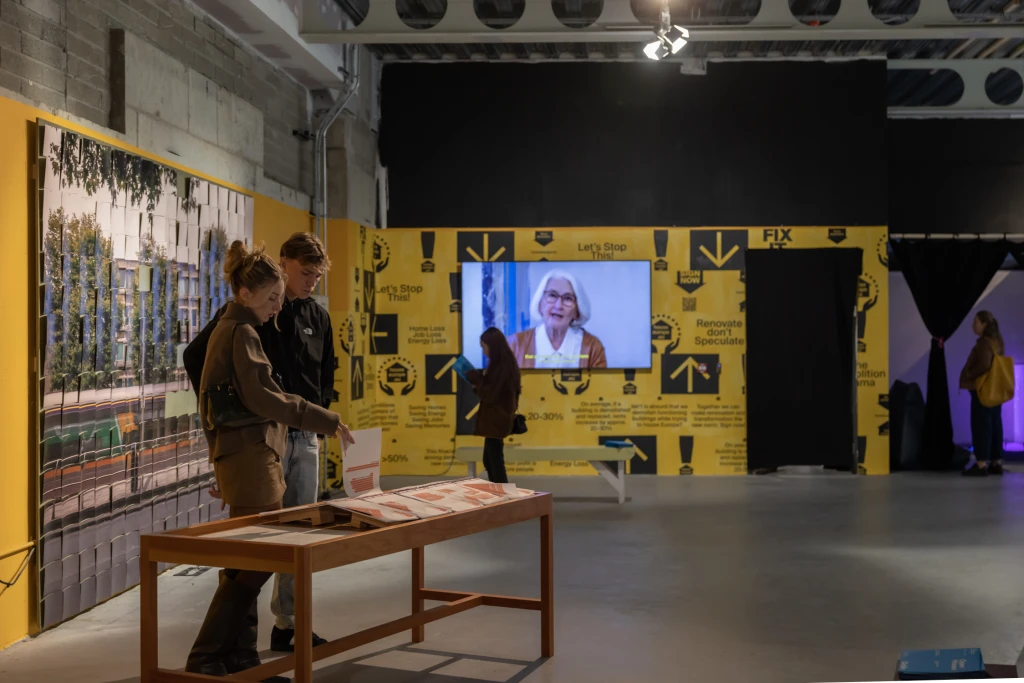 A part of the exhibition space where two bright yellow walls meet in a corner in the background. The wall on the right has a flat screen playing a film, and a visitor is looking at it and the facts and slogans posted on the wall. The left wall has a huge photo of O'Connell Street. In front of it, in the foreground, is an open topped display case. A young man and young woman are looking down and flipping through the material in the case.