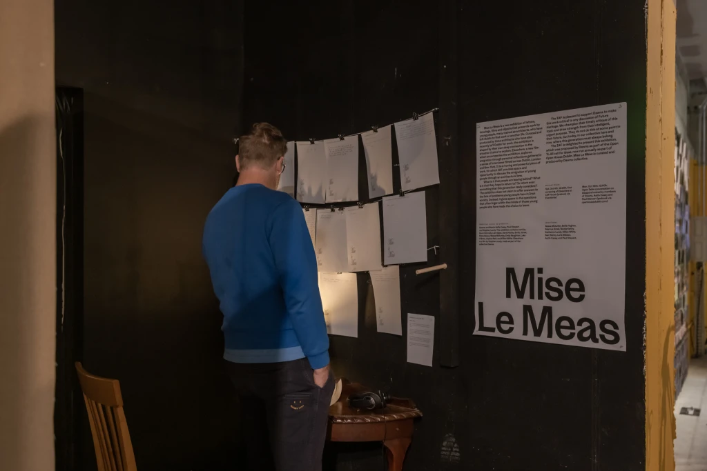 A man wearing a blue top and black trousers stands at a writing desk and chair. On the wall above the desk is a string with pieces of paper hanging from it - notes and drawings by visitors. The exhibition text, titled Mise Le Meas, is on the wall to the right of the desk.