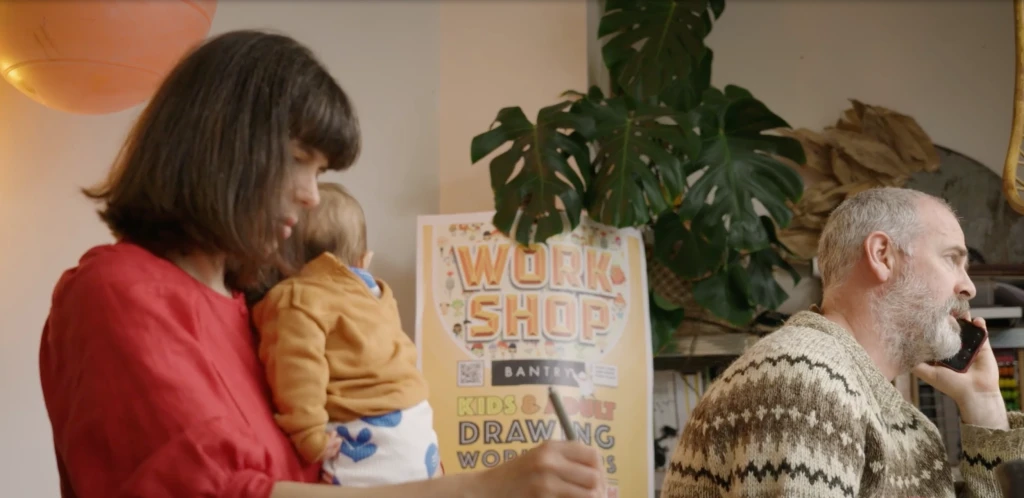 Inside a studio space, a woman holds a baby with her left arm and draws with her right hand. A man is on a mobile phone. In the background is a sign for Work Shop Bantry.