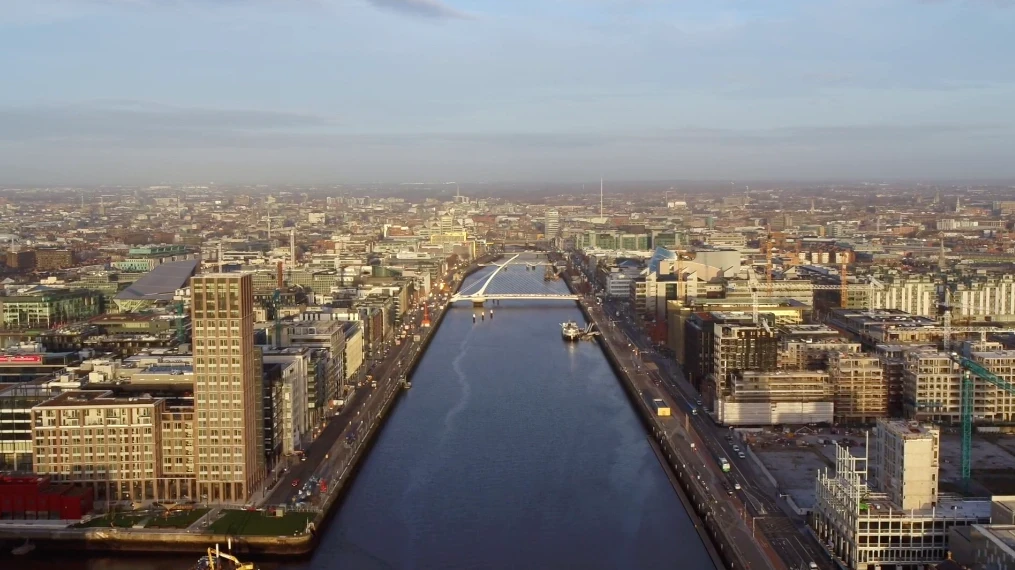 Aerial shot of Dublin looking down the Liffey with the harp-shaped Samuel Beckett bridge, boats, and tall buildings either side. The water is dark blue while the sky is a lighter, hazy blue with purple on the horizon.