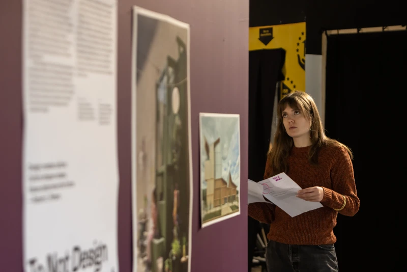A shot along a purple internal wall on which the exhibition intro text and two photos are displayed. A young woman is looking up at the display around the corner and holding a printout of the catalogue.
