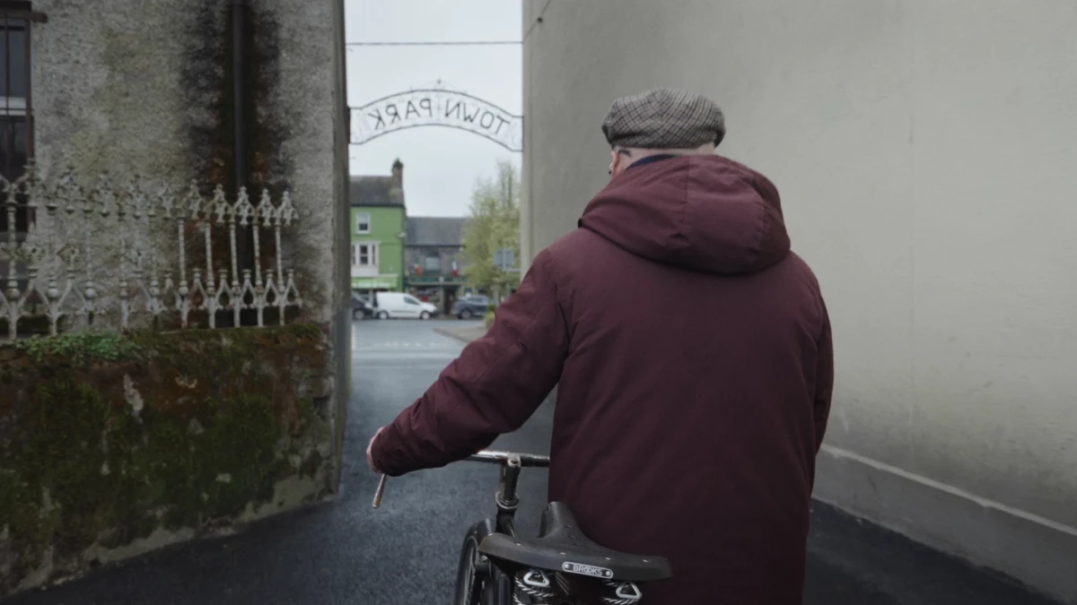 An older man wearing a winter coat and flat cap is pushing a bicycle on a path that narrows between two buildings. An arch-shaped sign over the path says Town Park.