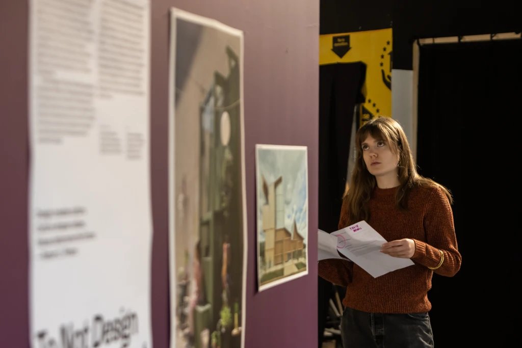 A young woman facing the camera is holding a sheet of paper and looking over it at the exhibition mounted on a dark purple wall in the foreground.