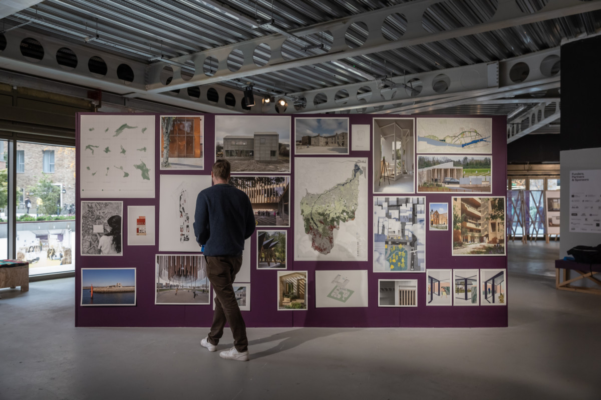 A man facing away from the camera is walking towards and looking at large photos and drawings mounted on a dark purple wall.