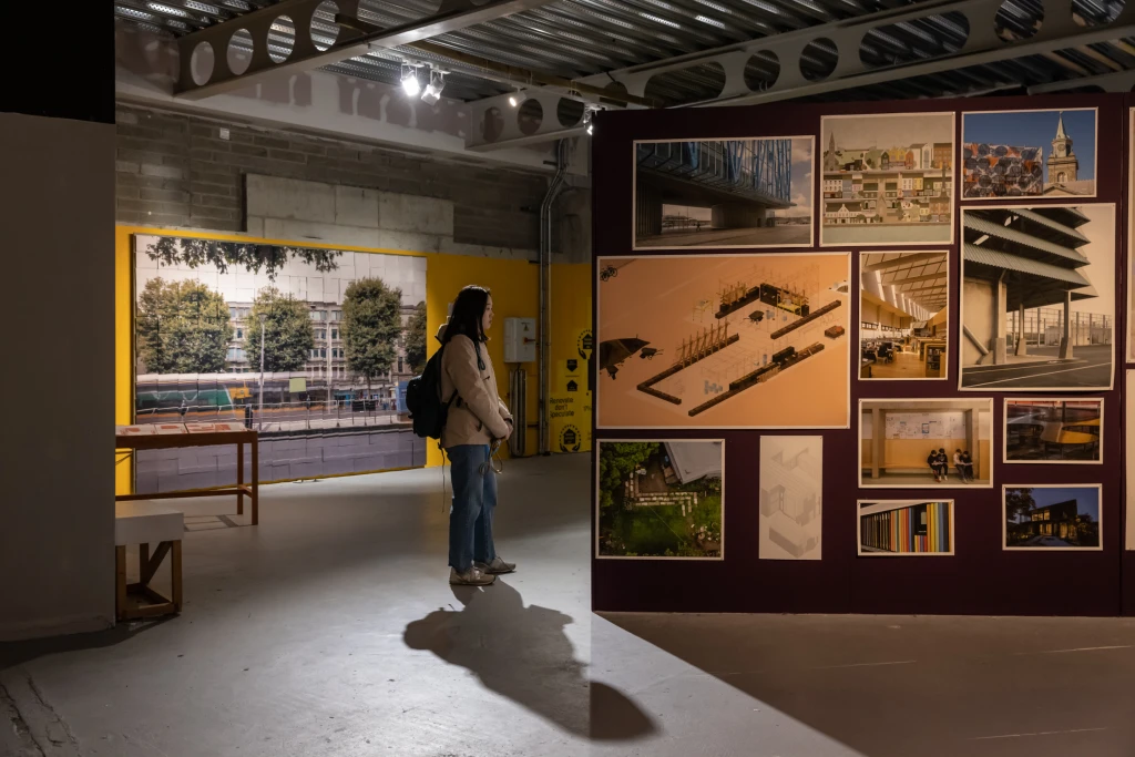 A woman with a backpack stands in the middle of the shot, with large photos and drawings mounted on the bright yellow wall behind her and on the dark purple wall to the right. She is looking at the display around the corner, unseen in the photo.