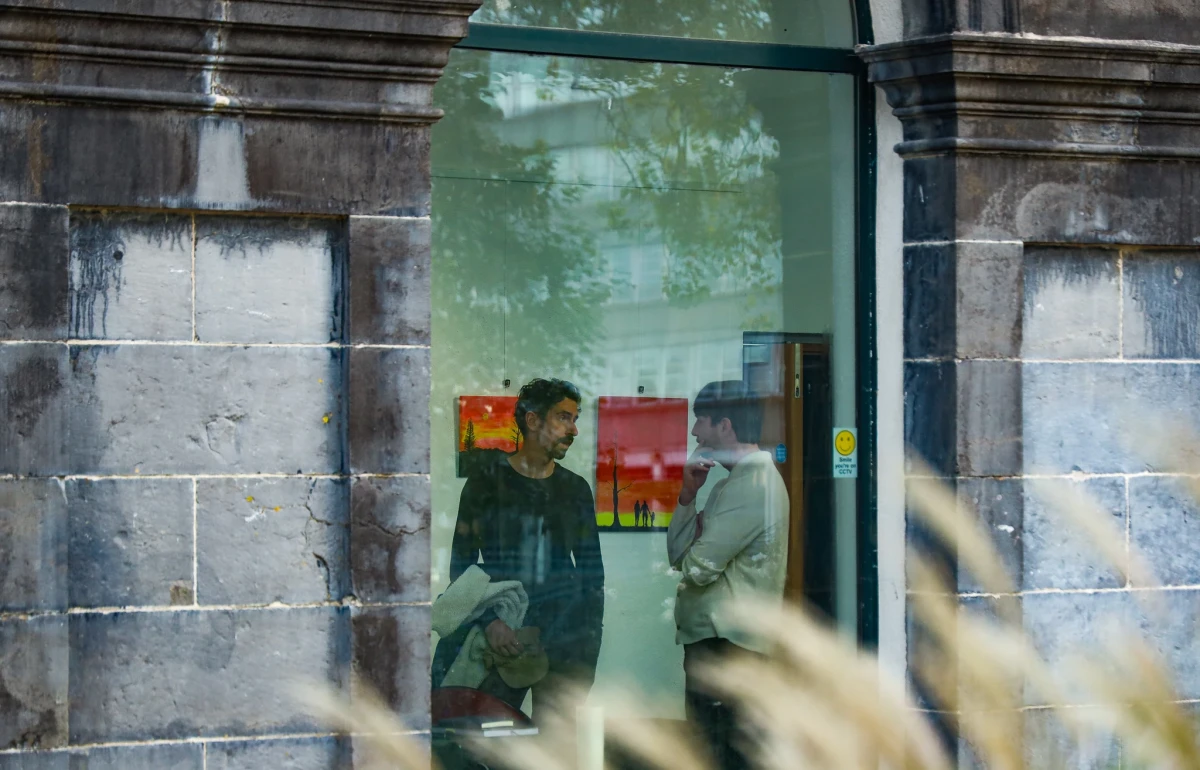 Exterior shot of a fancy stone building and looking in through the window at two men talking in a white room with paintings on the wall behind them. The window reflects the trees outside, and there are blurry ornamental grasses in the foreground.