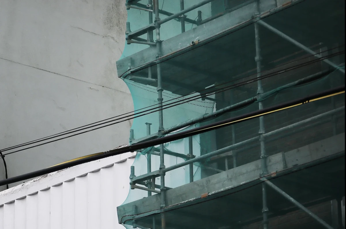 Looking up at a building wrapped in scaffolding and a green protective net, which contrasts with the grey and white building beside it.