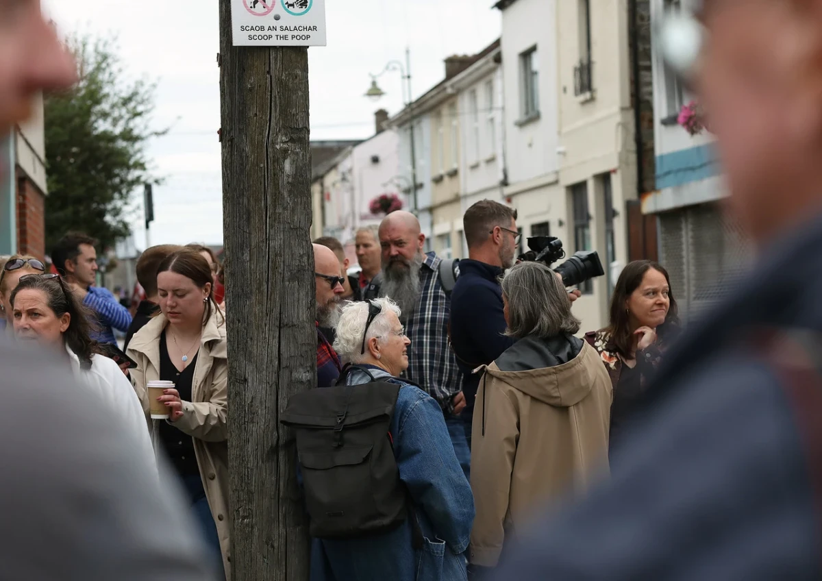 A group of people wearing autumn jackets gathered on a street, seen betwen the shoulders and heads of two men in blurred focus in the foreground. Some of the people are leaning against a wooden electricity pole. There are two-storey buildings in the background.