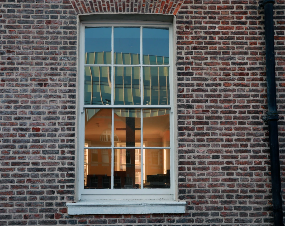 A white sash window in a Georgian red brick wall reflects a modern green roof from across the street. Through the window, the interior is a warm peach colour.