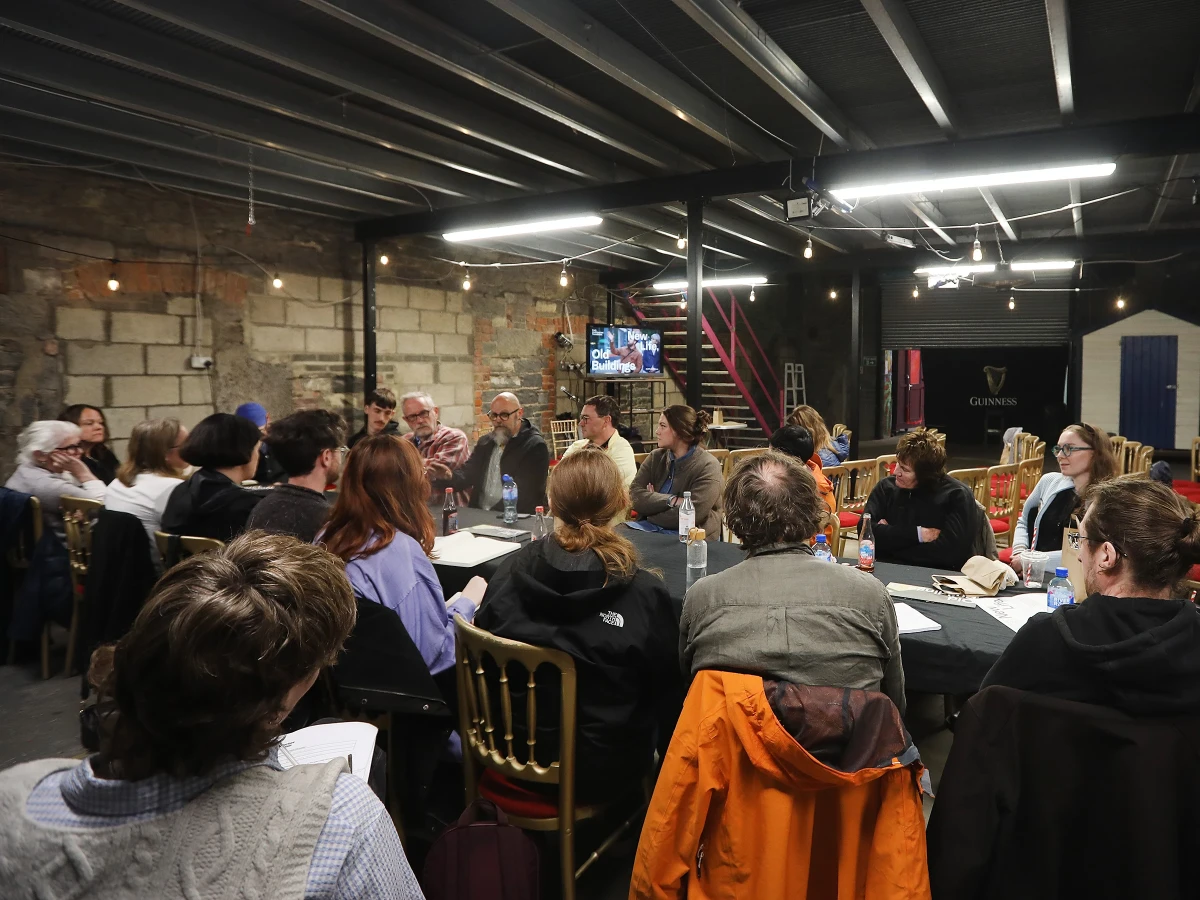 About 20 people having a discussion around a long table with a black tablecloth, in a semi-finished interior.