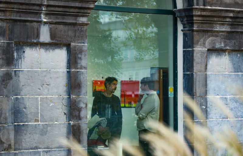 From outside, looking through a window in a stone building that has textured detailing. Inside, two men are talking in a room with art hanging on the wall behind them.