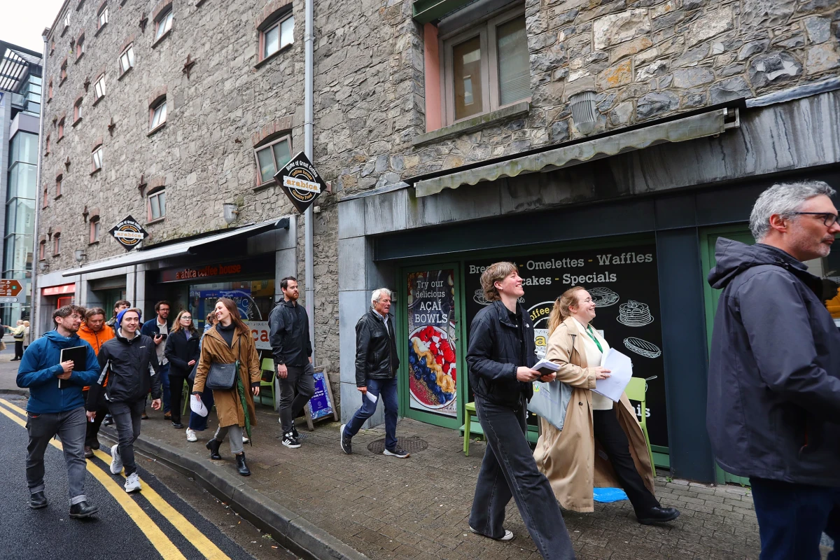 A group of people walking and chatting on the sidewalk in front of a tall stone building with small windows and with shopfronts at ground level.