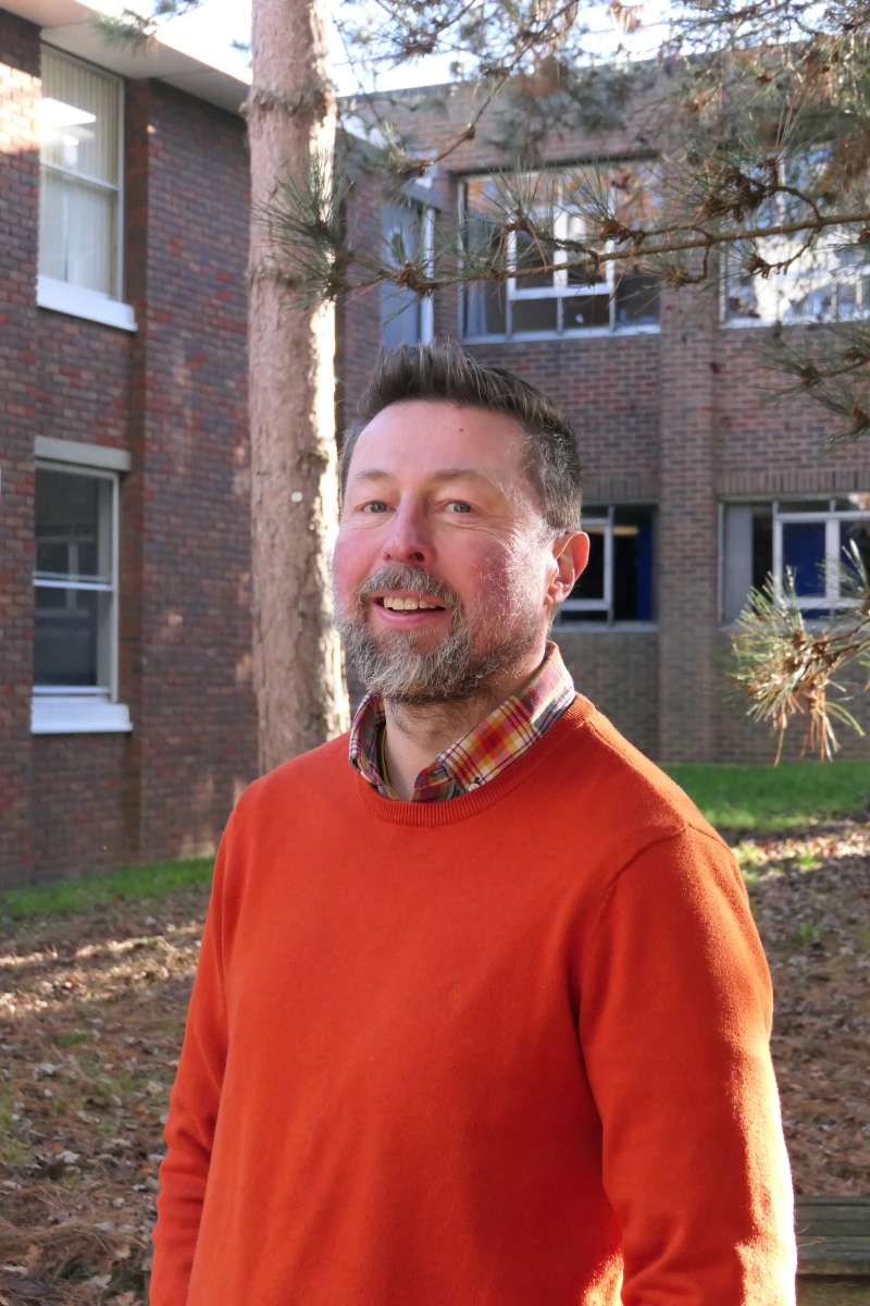 A man wearing an organge jumper stands in front of a brown brick apartment complex. He is smiling slightly.