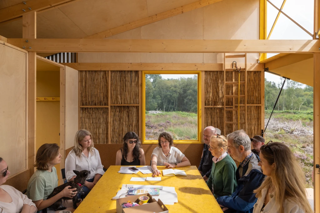 Interior shot of people of different ages sitting around a bright yellow table inside a cabin made of wood. There are writing materials on the table. The back wall of the cabin is lined with reeds and has a window opening onto the bog. The right of the cabin is completely open, with the wall panels raised to form an awning and lowered to form a platform.