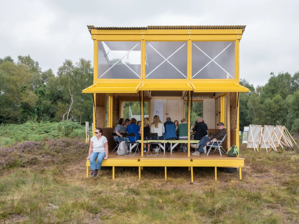 A group of people are sitting on benches and chairs around a table in a bright yellow cabin with big windows. The front of the cabin is open. A woman sits on the ledge at the front left corner of the cabin, facing out to look at the bog.