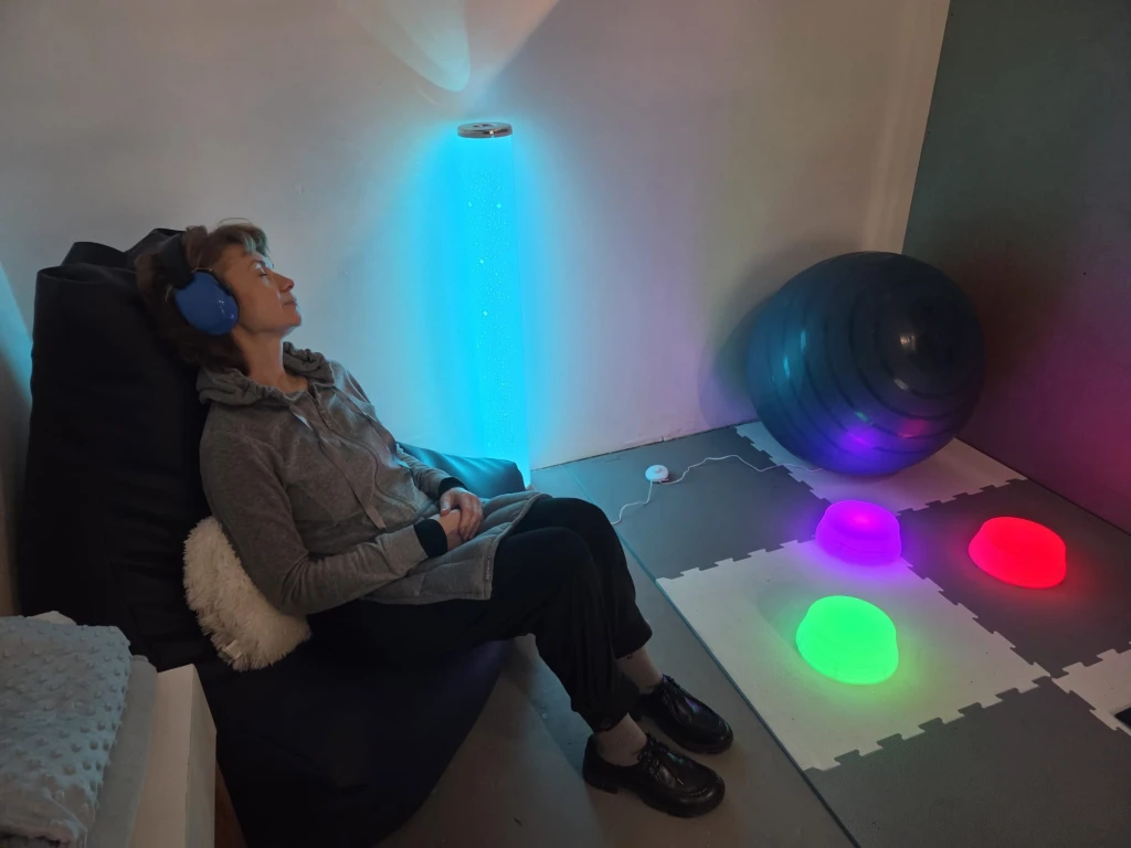 A woman reclines on a soft leather beanbag chair. She is wearing big noise cancelling headphones and her eyes are closed. There are coloured lights but the room is not too bright. There is a yoga ball and a stack of folded blankets.