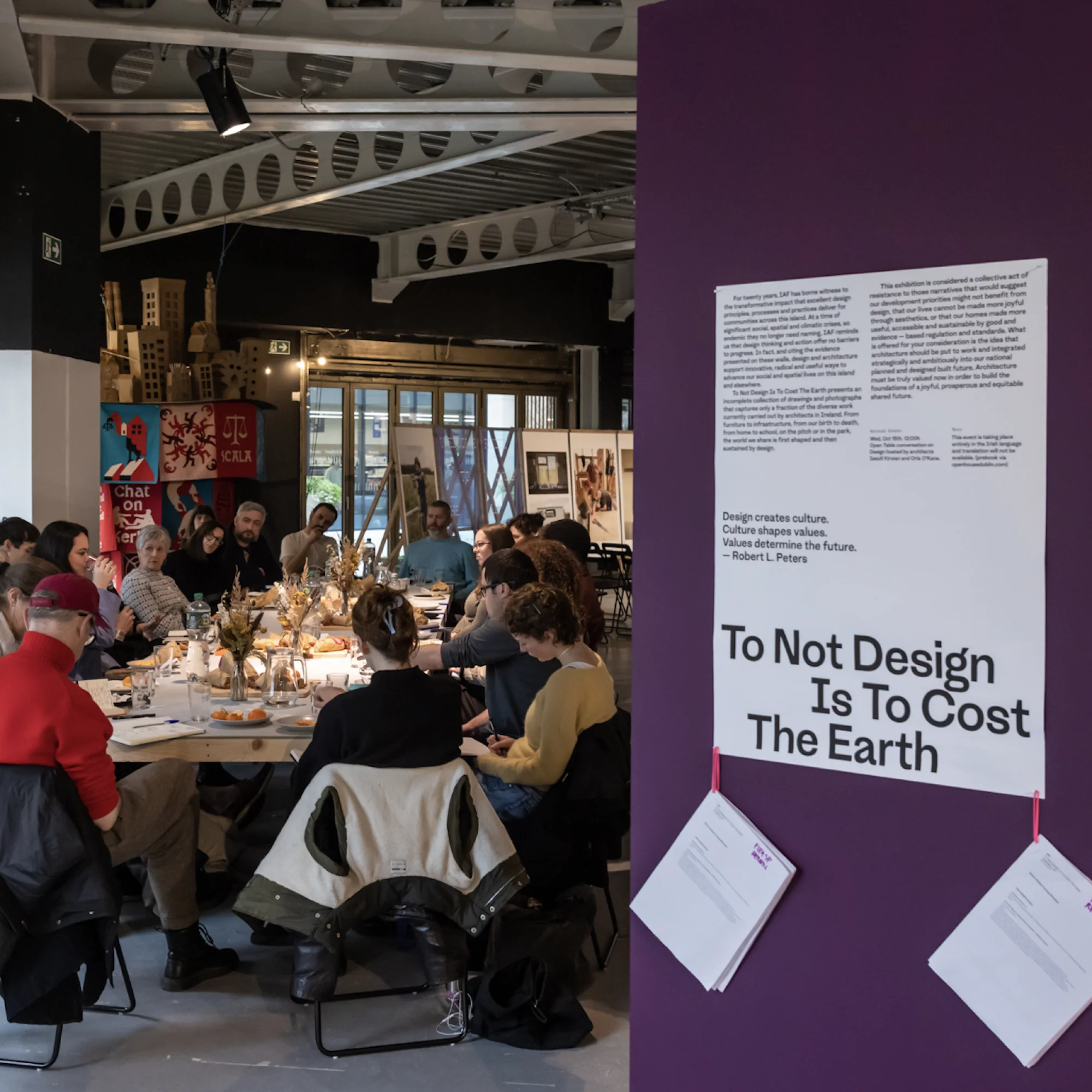 In a warehouse-like space, a group of people sit around a table with food, having a conversation. There is a glass wall to the left. To the right and in the background are exhibitions. The one to the right shows text and photos mounted on a dark purple wall.