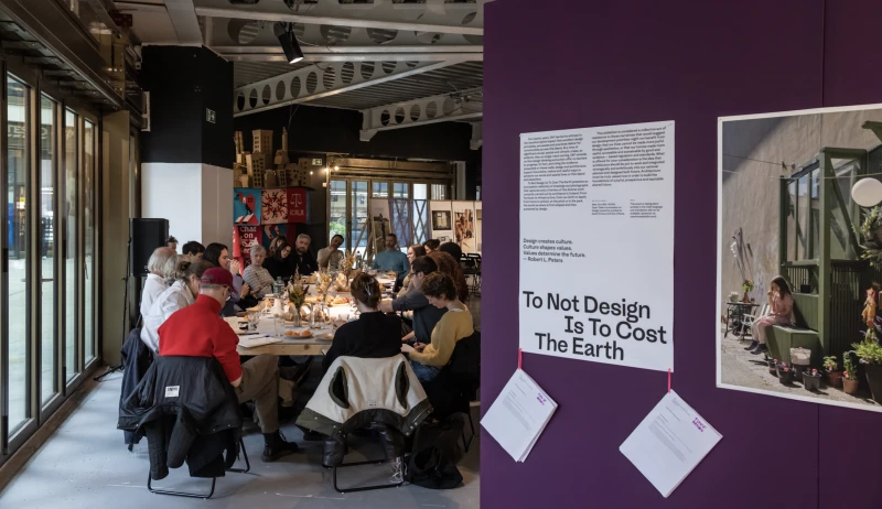 In a warehouse-like space, a group of people sit around a table with food, having a conversation. There is a glass wall to the left. To the right and in the background are exhibitions. The one to the right shows text and photos mounted on a dark purple wall.