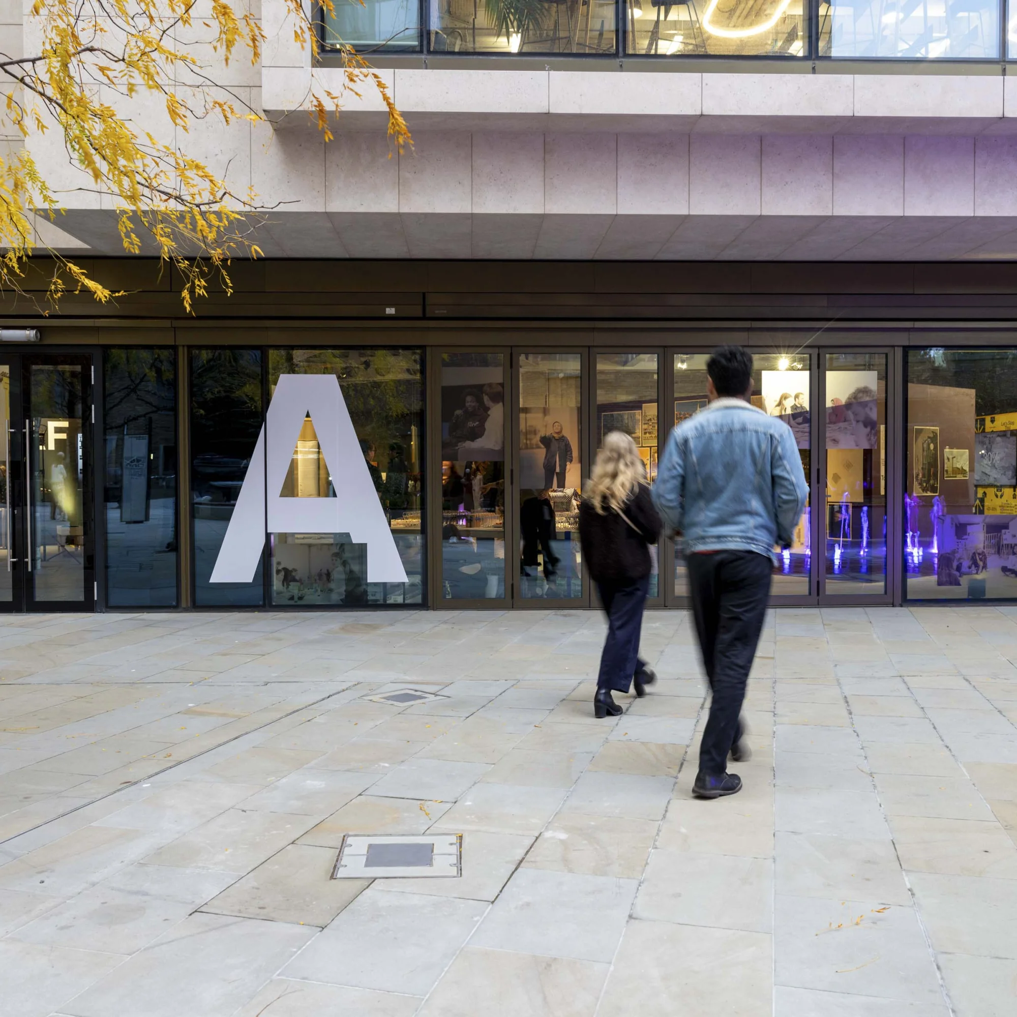 A young man and woman with long blonde hair seen from behind as they walk across a stone-paved square towards the door of a glass-fronted building. The letters IAF are on the glass.