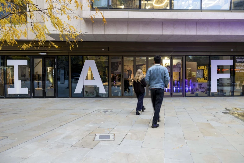 A young man and woman with long blonde hair seen from behind as they walk across a stone-paved square towards the door of a glass-fronted building. The letters IAF are on the glass.