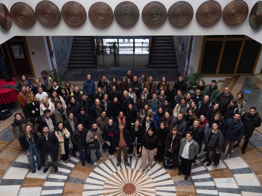 A large group shot, looking down on them from a balcony. The people are standing on a tiled floor with a sunburst pattern. Behind them are stairs and doors, and there is a white overhang decorated with large metal spirals hung like shields.
