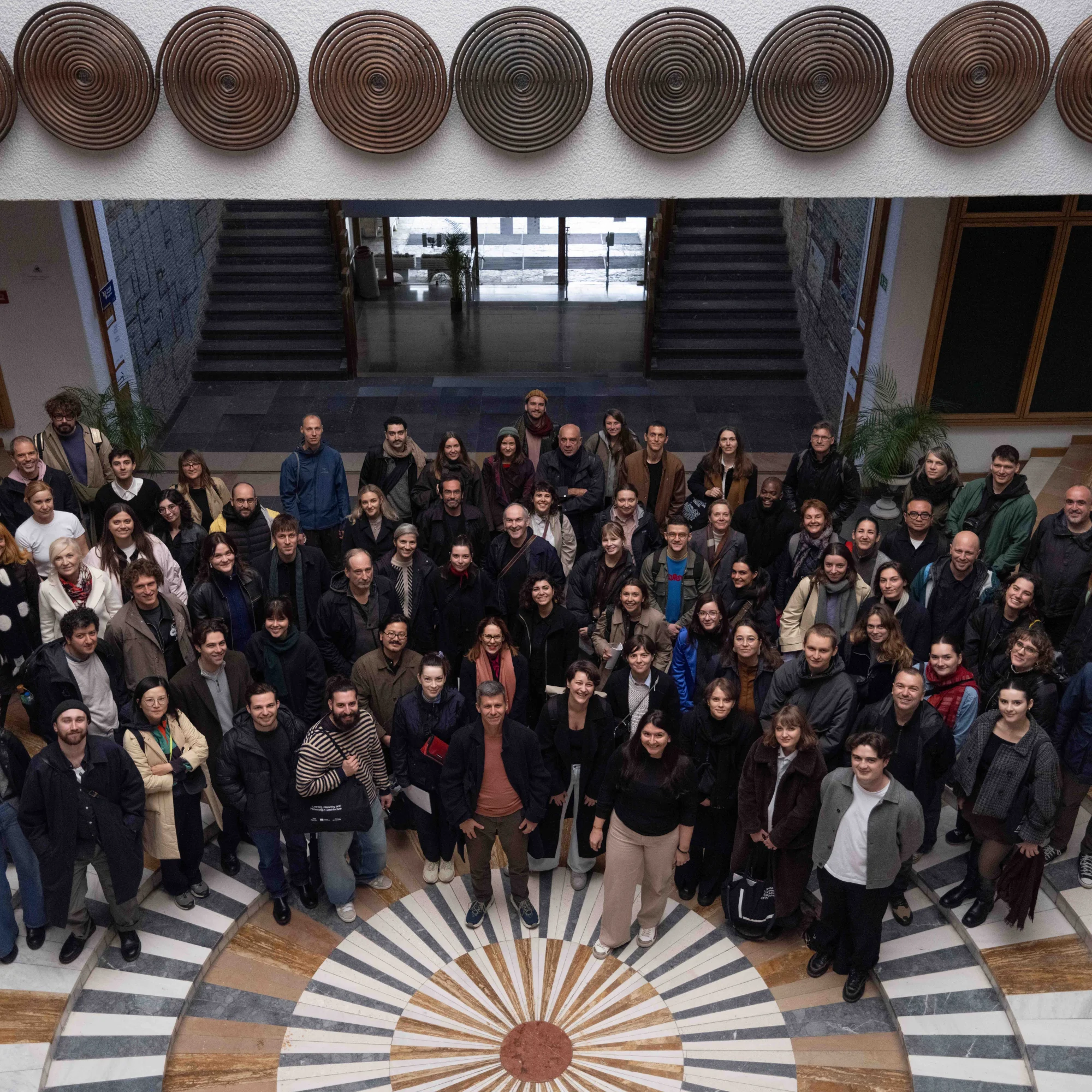 A large group shot, looking down on them from a balcony. The people are standing on a tiled floor with a sunburst pattern. Behind them are stairs and doors, and there is a white overhang decorated with large metal spirals hung like shields.