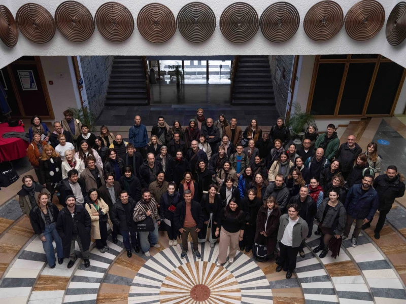 A large group shot, looking down on them from a balcony. The people are standing on a tiled floor with a sunburst pattern. Behind them are stairs and doors, and there is a white overhang decorated with large metal spirals hung like shields.
