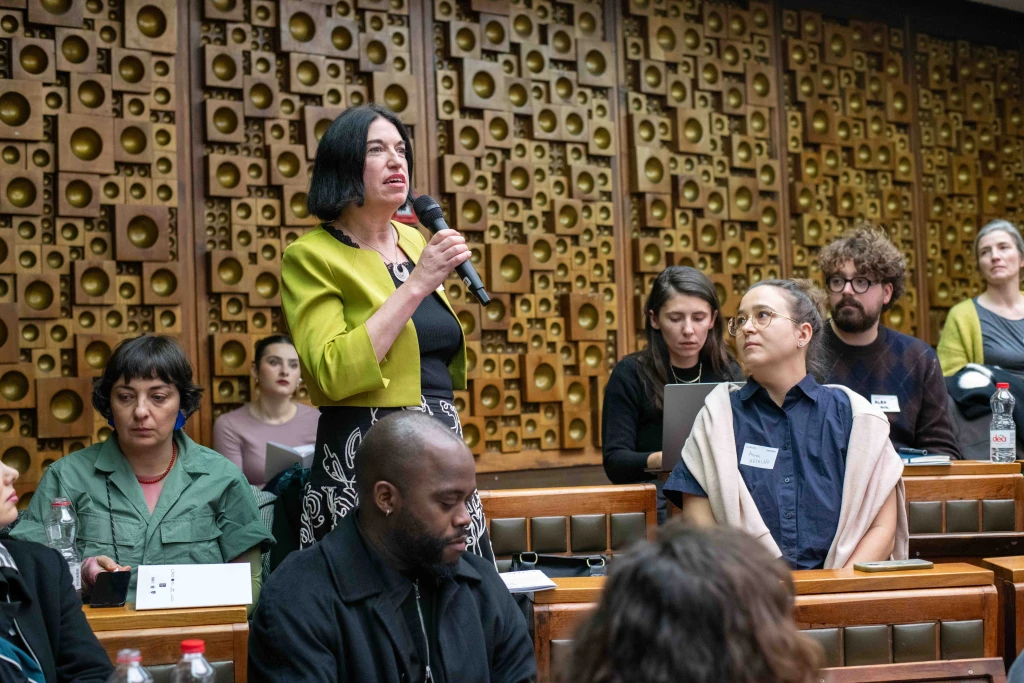 A white woman with dark hair, wearing a black dress with a white floral pattern and a bright chartreuse jacket, is standing and speaking into a mic. Other delegates are seated around her, in a room with fixed bench and desk seating and wood panelling made of a collage of wooden blocks with hollowed out circles in the middle.