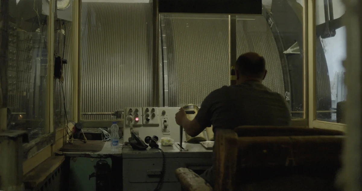 Colour photo from behind of a man sitting at a control board, with his left hand on a lever. In front of him is a large wheel on the other side of a glass wall.