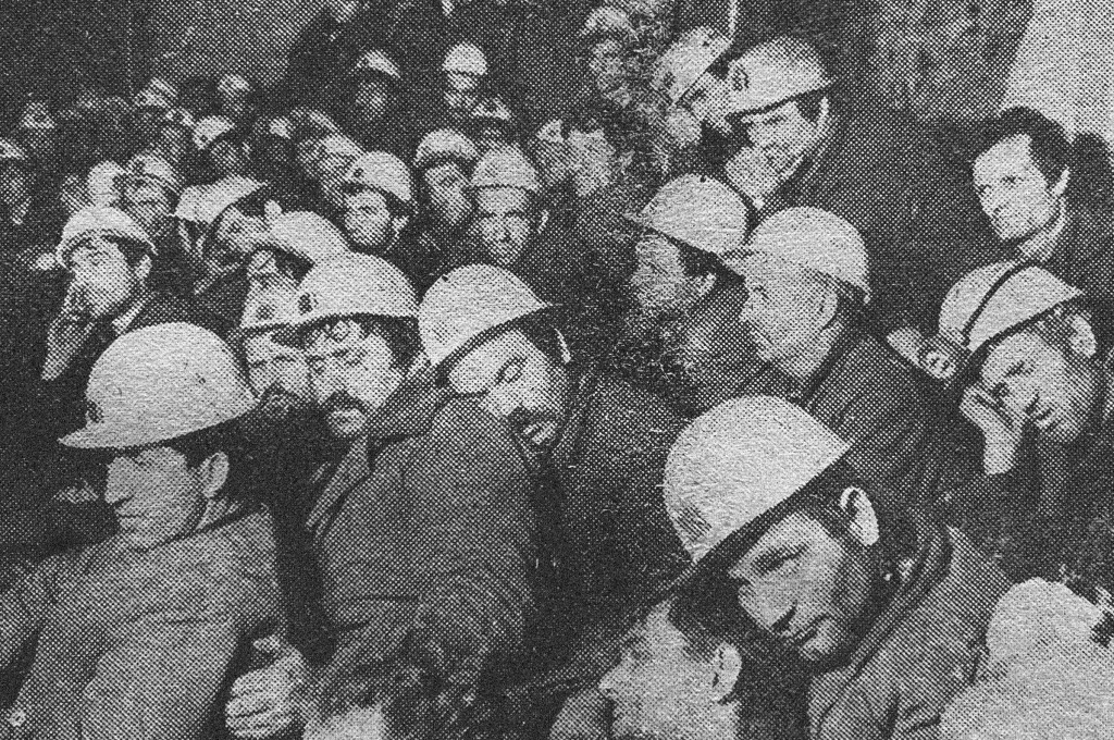 Grainy black and white photo of miners in their work clothes and hard hats, crowded together in the mine.