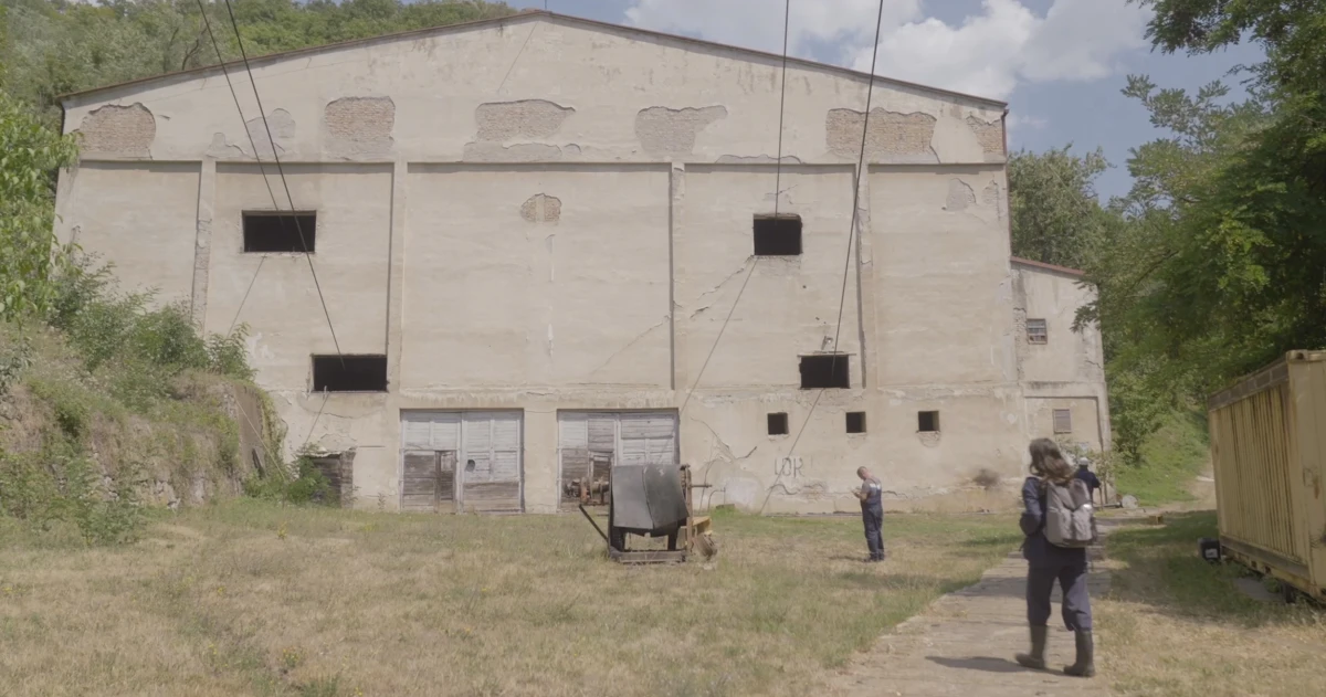 Colour photo of someone wearing a backpack approaching a large warehouse-like building with small windows and chipped paint. Cables run out of the windows overhead. The grass in front of the building is scorched and there are trees and undergrowth around the building. Someone else stands between the building and the person approaching.
