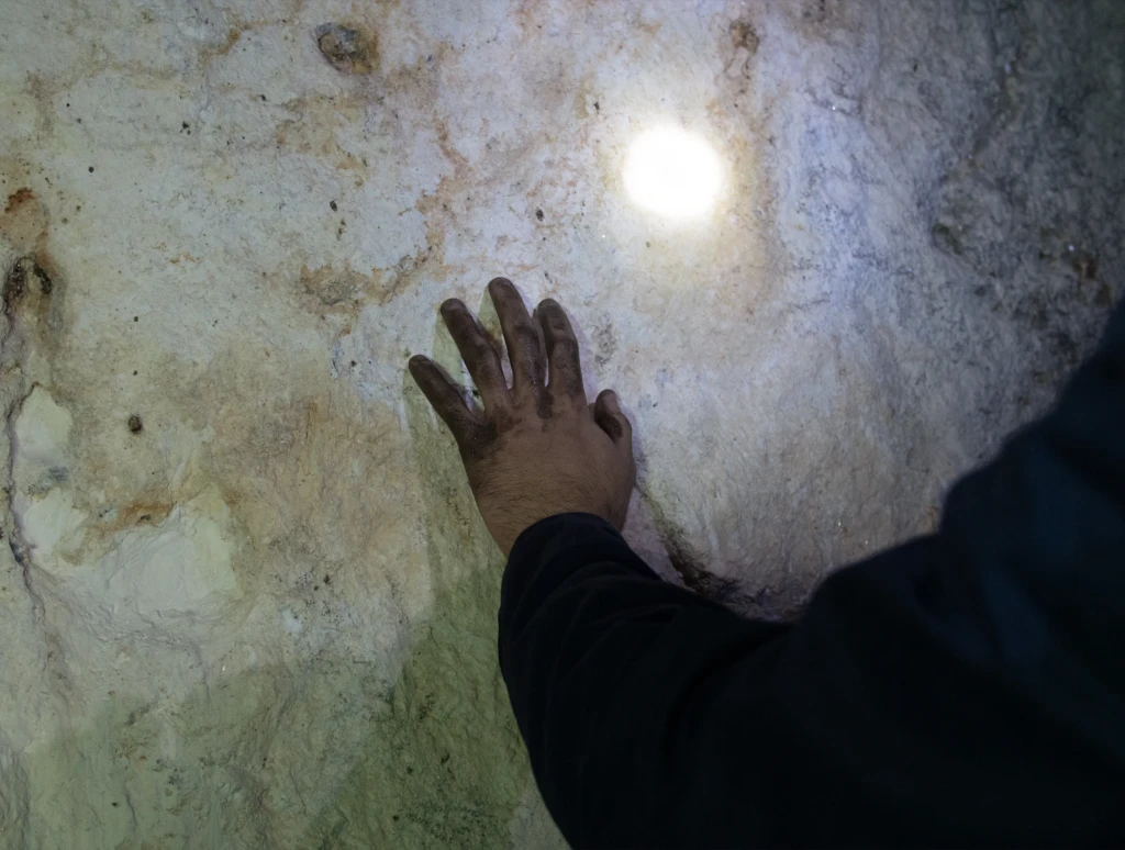 A dirty hand feeling the uneven surface of a white wall with a vein of minerals. There is a bright light shining on the wall, probably from a headlamp.