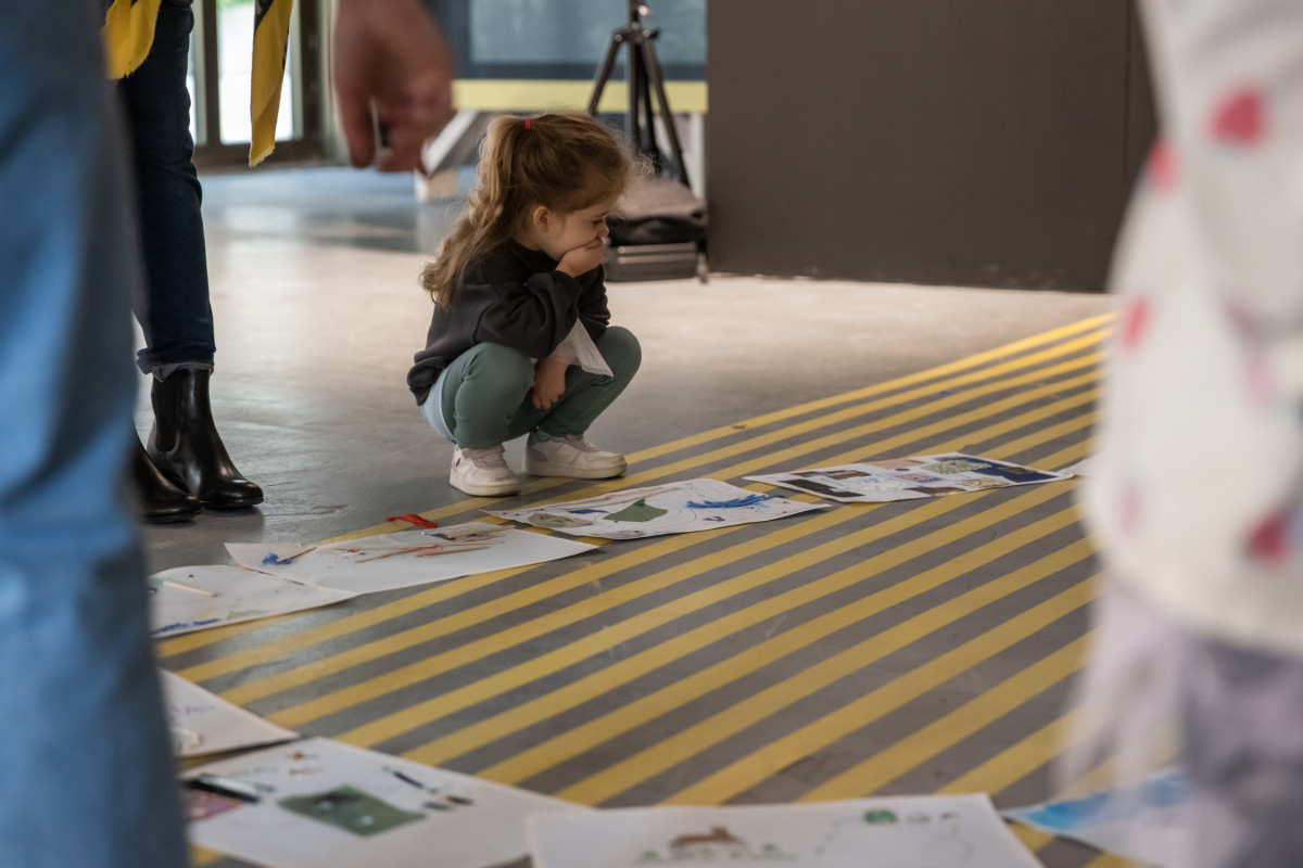 A young girl with a long ponytail squats on a yellow-striped floor, looking at children's drawings and collages laid out in a circle.