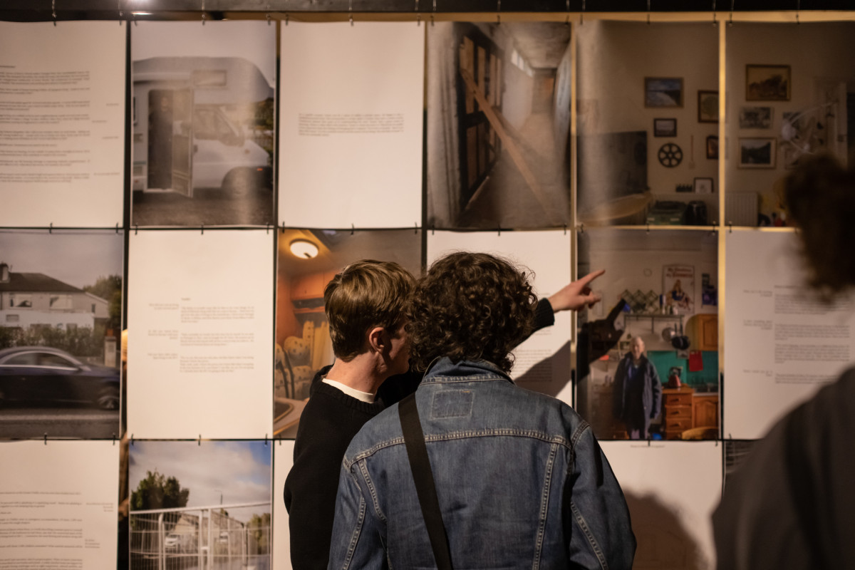 Two young men looking at a display of photos and texts that hang vertically. The photos show squashed living conditions. One of the young men is pointing at one of the photos.