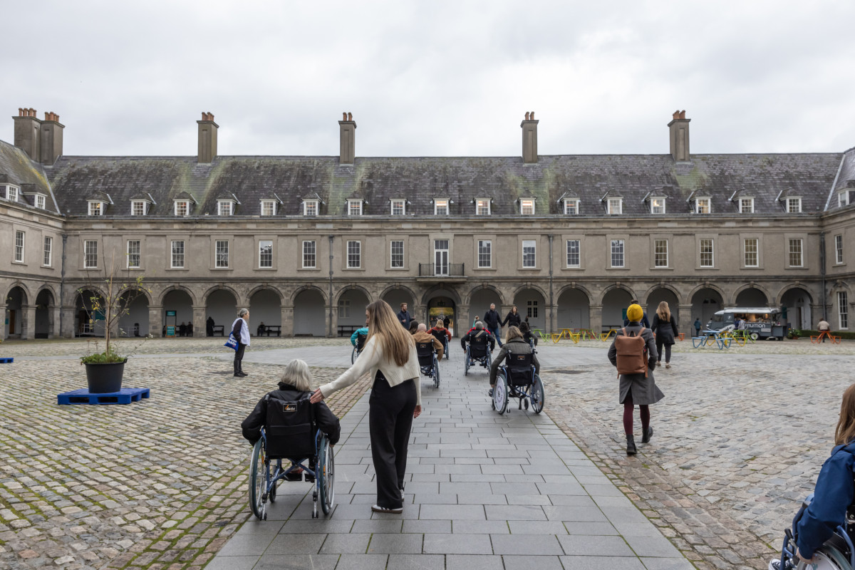 Seen from behind, several people are crossing a large, cobbled courtyard. Some are in wheelchairs and others are walking. There is a central path with flat paving stones but the rest of the surface is very uneven. There are some potted plants in the courtyard. The surrounding historic buildings are made of stone, with rows of arches, windows and chimneys.