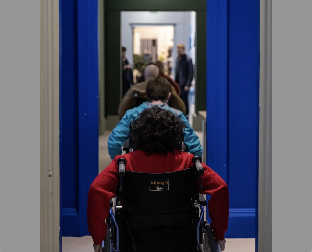 Seen from behind, a row of people in wheelchairs are navigating through a series of doorways inside a gallery.