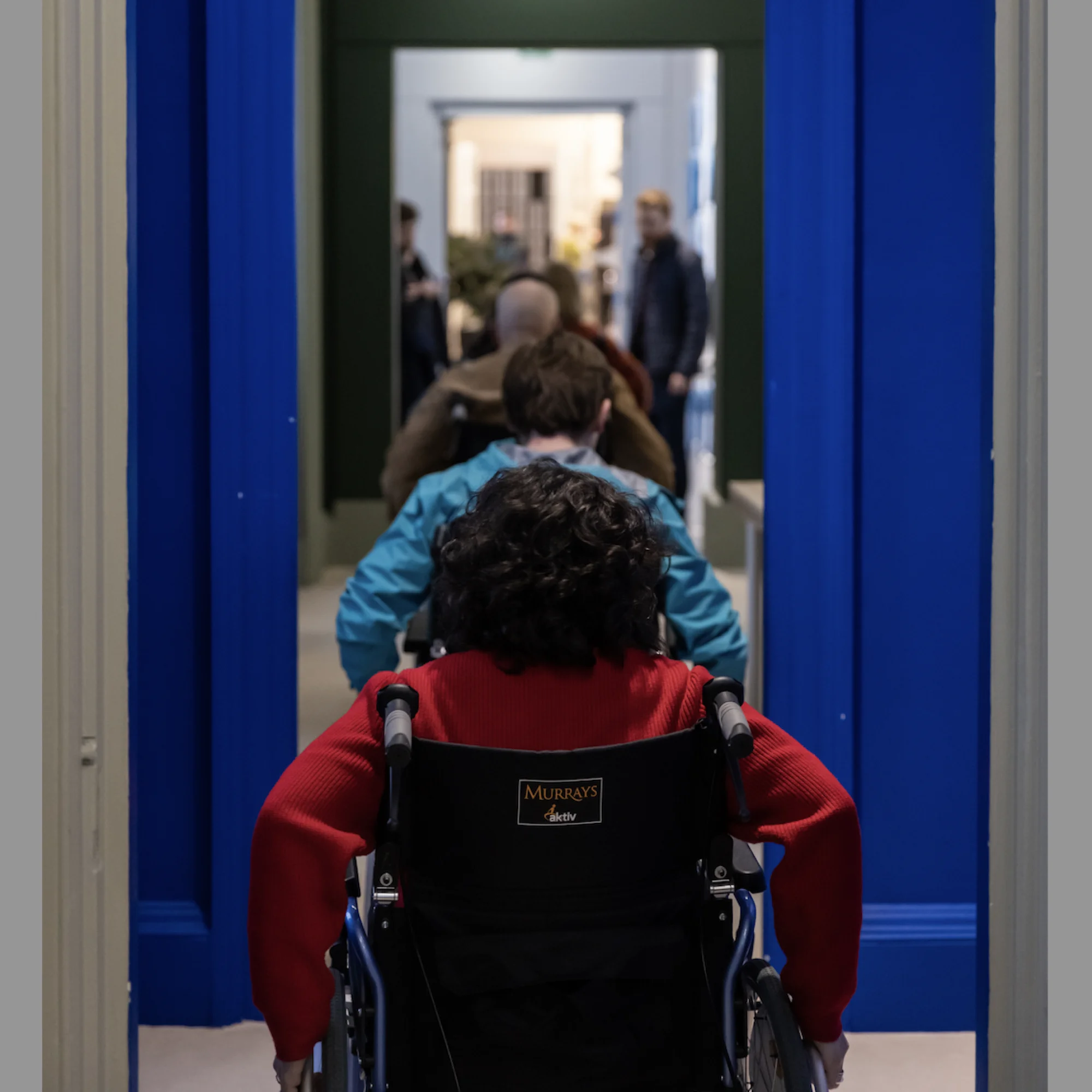 Seen from behind, a row of people in wheelchairs are navigating through a series of doorways inside a gallery.