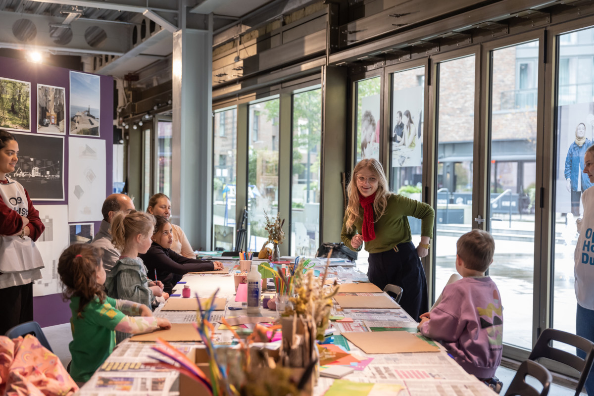 Young children and parents sit around a long table covered with craft supplies. An Open House Dublin volunteer stands to the left, while the woman leading the workshop stands to the right. She is smiling, speaking and gesturing to the children. The room has a display of images on a purple wall background left and a wall of windows background right.