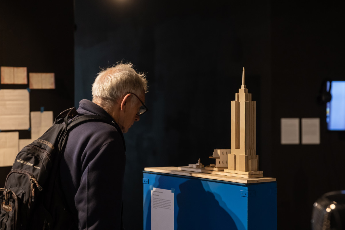An older man is looking at a model of the Empire State Building on a bright blue plinth. In the background, letters and a flatscreen TV are mounted on black walls.